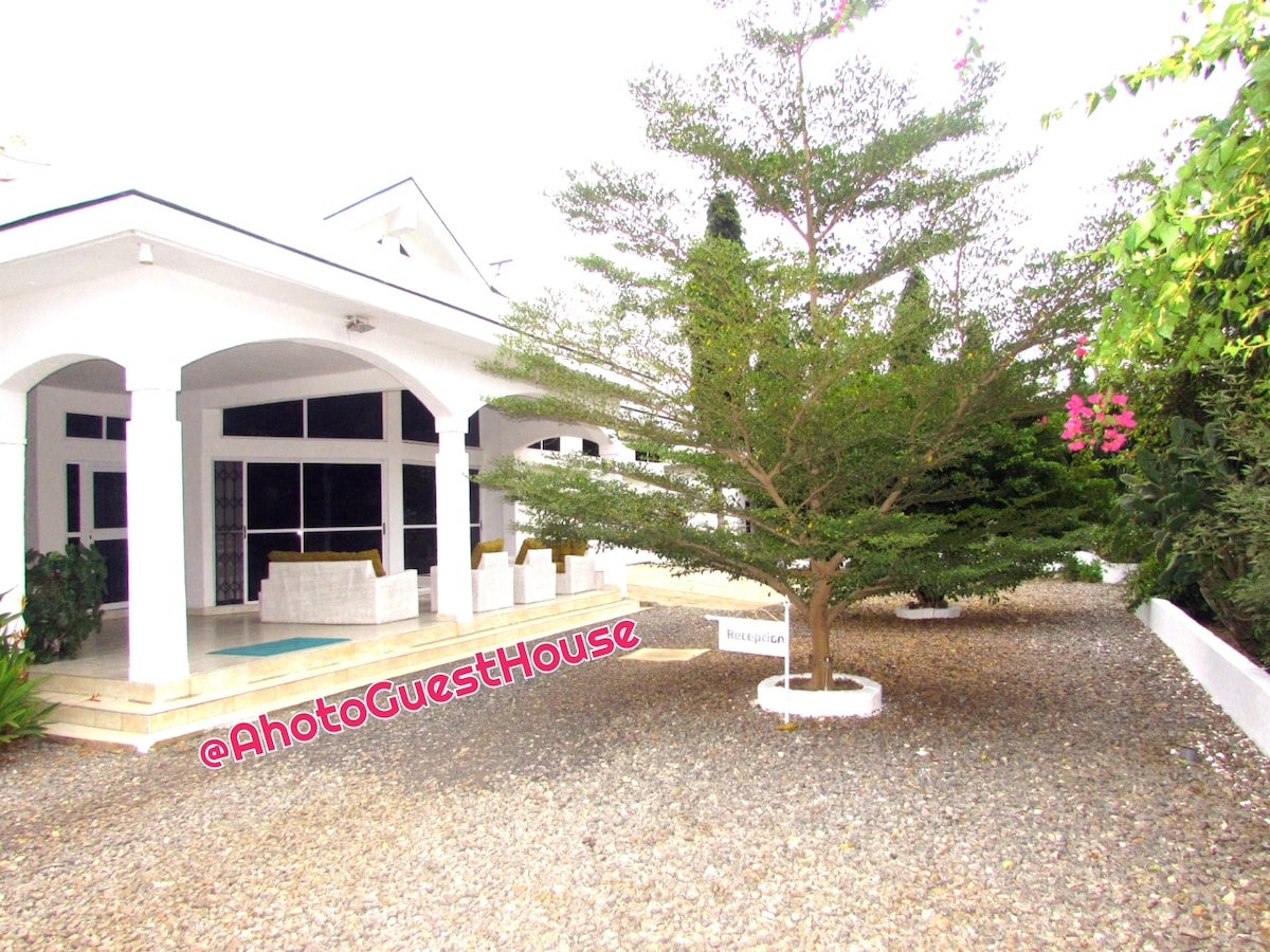 An elegant guest house exterior is framed by lush greenery and stone walkways. A sizable tree provides shade near the entrance, while the white building features large windows and a welcoming porch area. A gravel driveway leads up to the structure, enhancing the serene atmosphere.