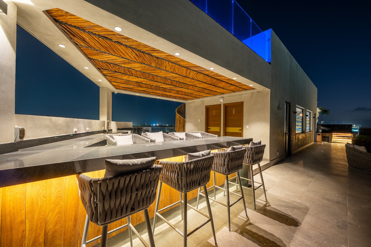 A modern rooftop terrace features a sleek bar with ample seating. The wooden ceiling adds warmth, contrasting with the smooth stone countertops. Soft lighting highlights the area, making it ideal for evening gatherings while providing open views of the surrounding skyline.