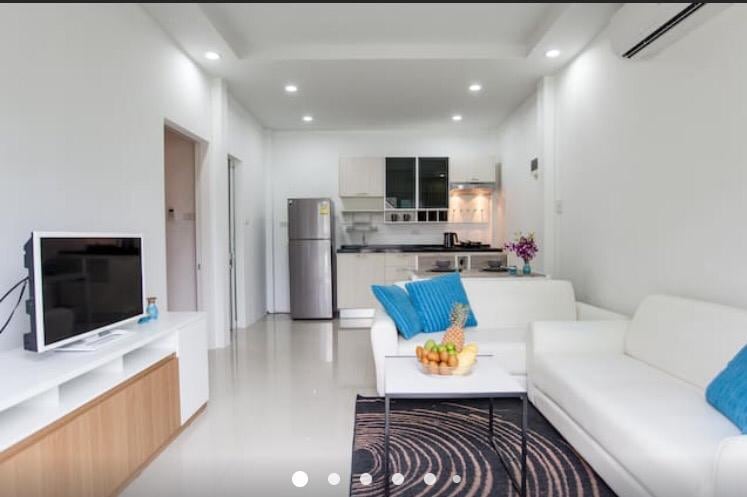 A modern living area is depicted, featuring a white sectional sofa with bright blue accent pillows. A sleek coffee table holds a bowl of fresh fruit. The open kitchen area showcases contemporary cabinetry and stainless-steel appliances, contributing to the space's clean and inviting aesthetic.