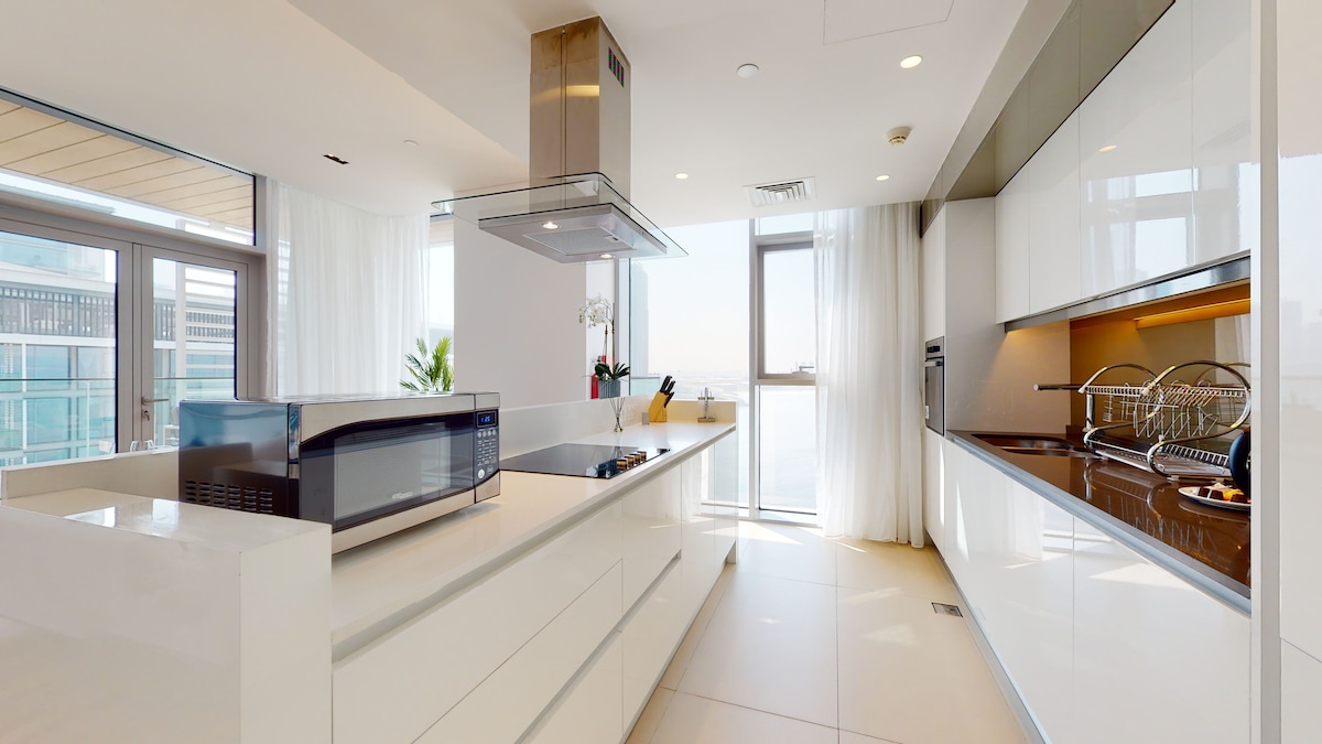 An open plan kitchen presents sleek white cabinetry and modern appliances. A large window allows natural light to flow in, offering views of the surroundings. A stylish kitchen island features a microwave and ample counter space, creating an inviting atmosphere for cooking and dining.