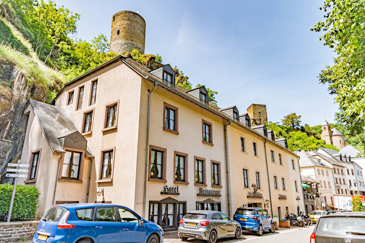 The exterior of Hotel Le Postillon is depicted, featuring a three-story building with a light-colored facade. Several vehicles are parked in front, and the ancient castle tower is visible in the background, surrounded by lush greenery and a clear blue sky.