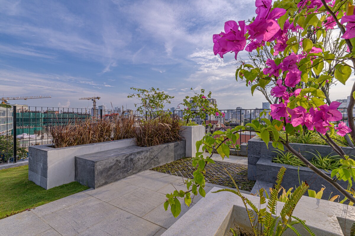 A serene outdoor space showcases vibrant pink bougainvillea flowers against a backdrop of soft green foliage. Modern benches made of stone are placed along the well-maintained pathway, providing a peaceful area to relax. The city skyline is visible in the distance under a clear blue sky.