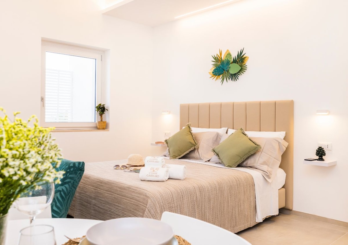 A serene bedroom features a spacious bed with neutral bedding and decorative green cushions. Natural light filters through a nearby window, illuminating the room. A simple wall decoration of leaves adds a touch of nature, while a small plant rests on the windowsill.