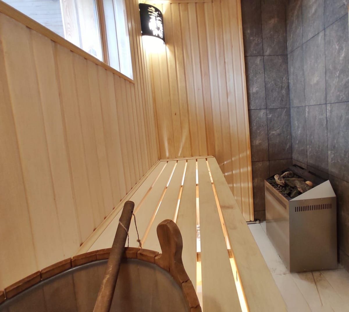 A wooden sauna interior is shown, featuring a slatted wooden bench and a stone-lined wall. A stove with visible stones is positioned in one corner, and natural light enters from a window, creating a serene environment for relaxation.