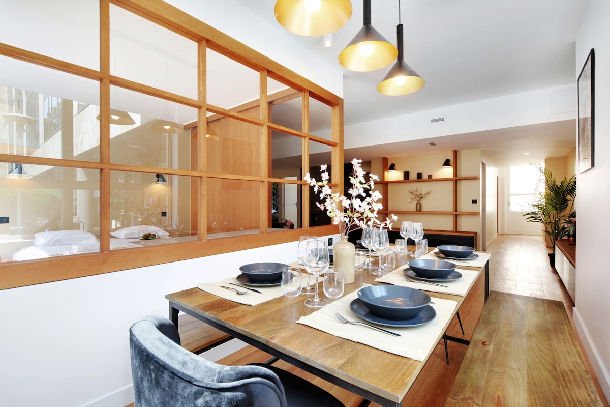 An open dining area features a wooden table set for six, adorned with plates and glasses. A light wood partition allows a view into the adjacent space, where soft lighting from hanging fixtures creates a calming ambiance.