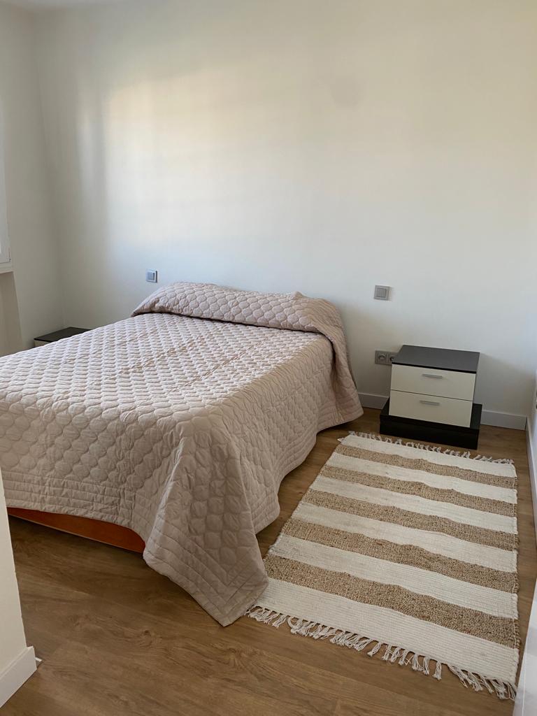 A well-lit bedroom features a spacious bed covered with a light quilt. A bedside table with drawers is located next to the bed. A textured area rug with alternating stripes is placed on the wooden floor, enhancing the room's neutral aesthetic.
