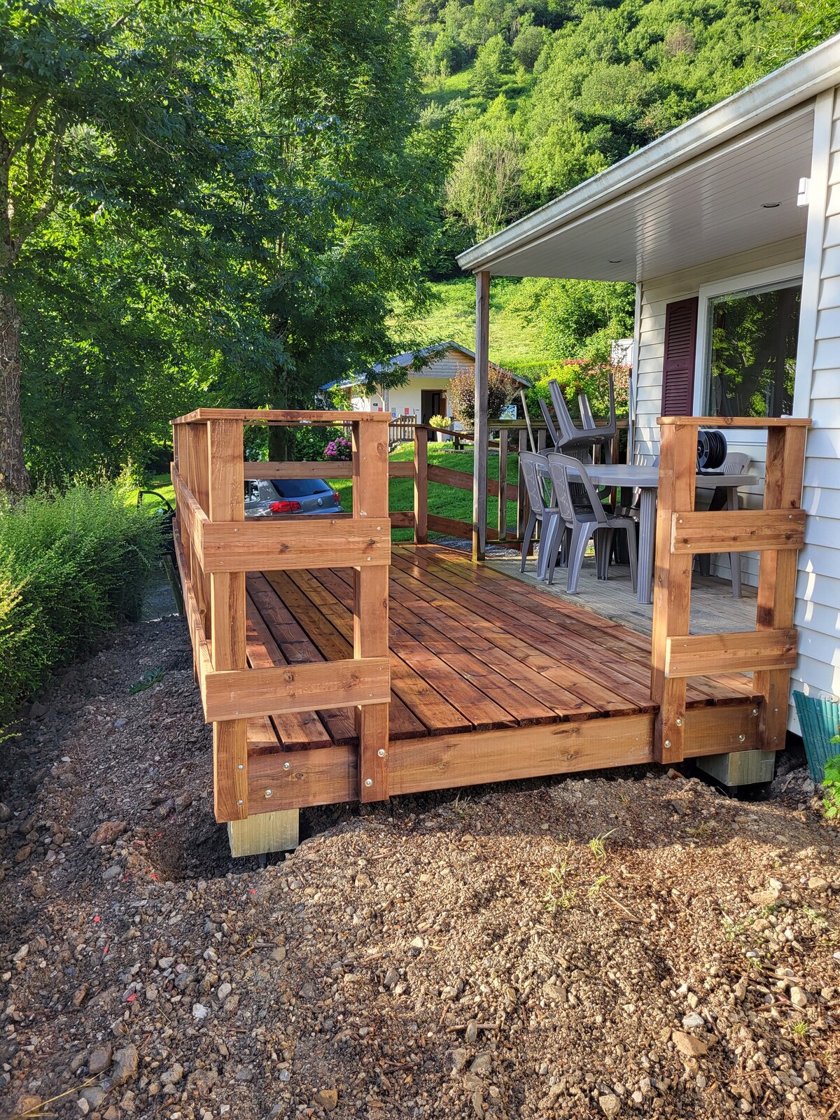 A wooden deck is positioned outside the mobil home, featuring a smooth surface and sturdy railings. A few chairs are arranged nearby. Lush greenery surrounds the area, and a view of the mountains can be glimpsed in the background.