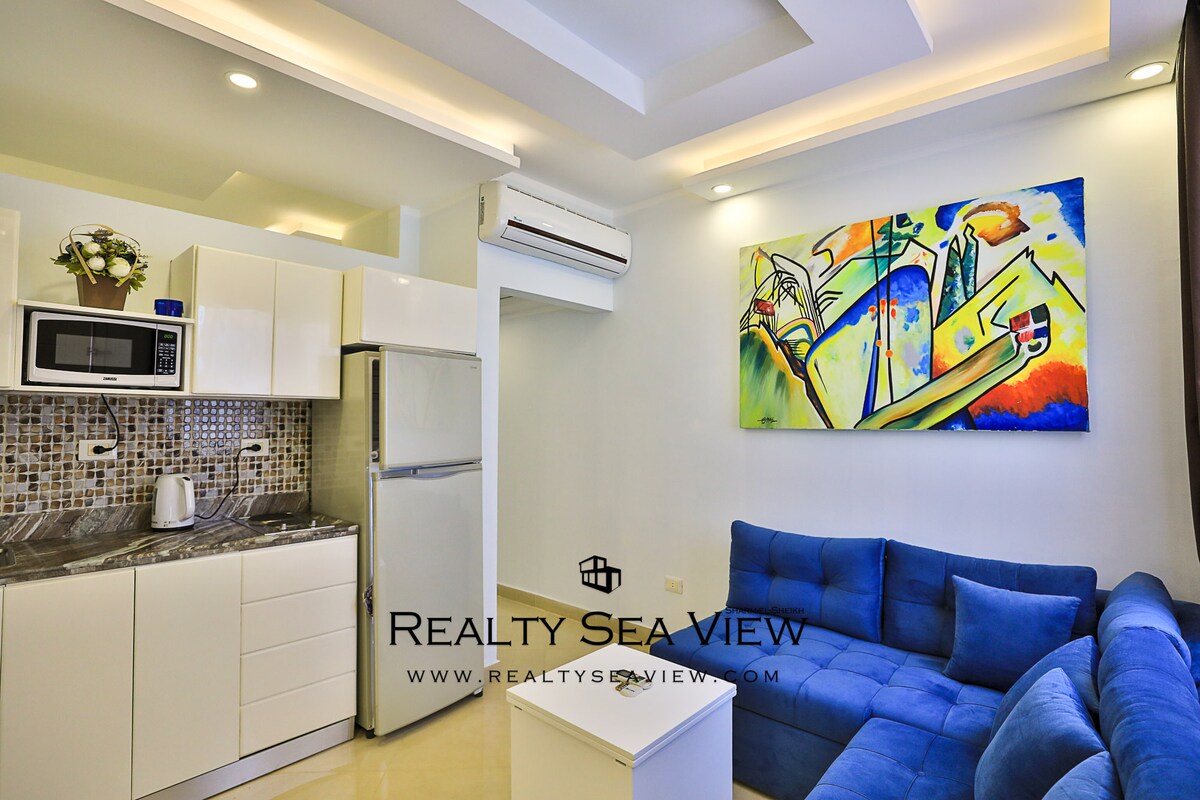 A modern kitchen area is visible, featuring sleek white cabinets and a compact microwave. A cozy blue sofa offers seating in the open living space. An abstract artwork adorns the wall, adding a splash of color to the room.