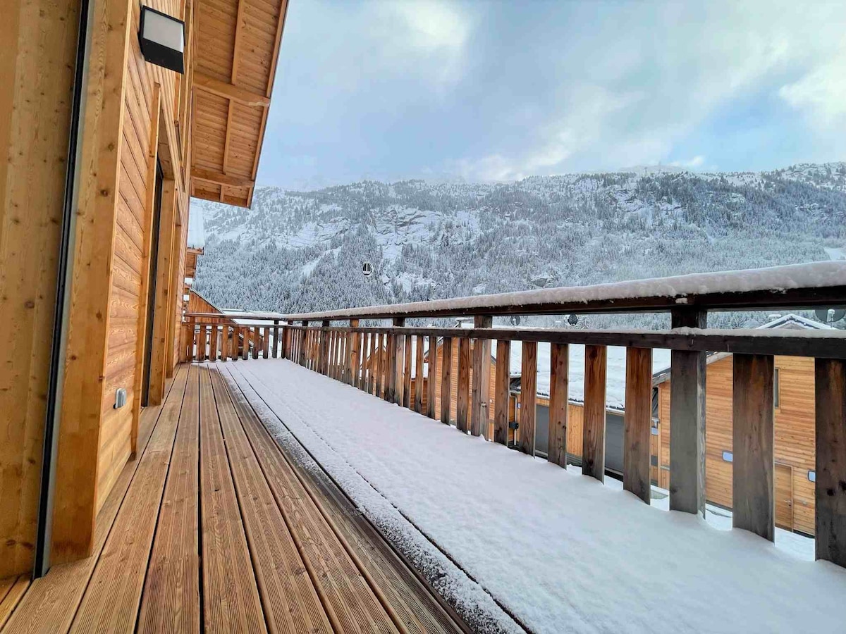 A spacious wooden balcony is visible, covered with a light layer of snow, extending towards the snow-capped mountains in the distance. The architectural design features wooden railings and walls, creating a warm contrast against the frost-covered landscape.