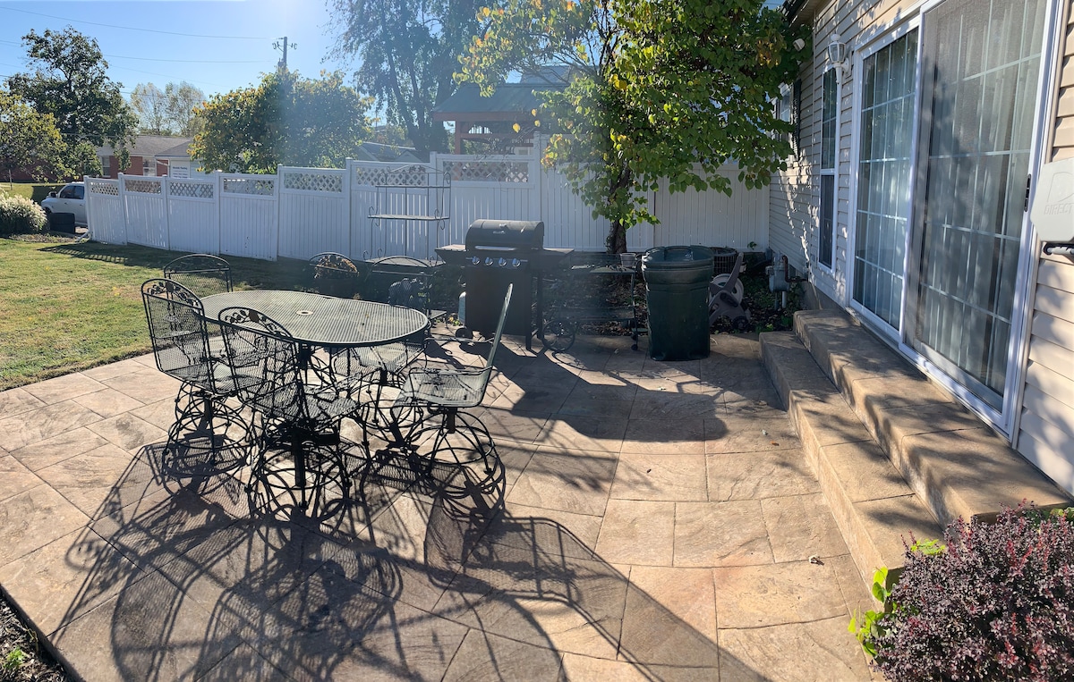 An outdoor patio area features a round table surrounded by six wrought iron chairs. A grill is positioned nearby, with a grass lawn and garden visible. Sunlight casts shadows on the stone surface, enhancing the peaceful setting and offering a welcoming space for outdoor dining.
