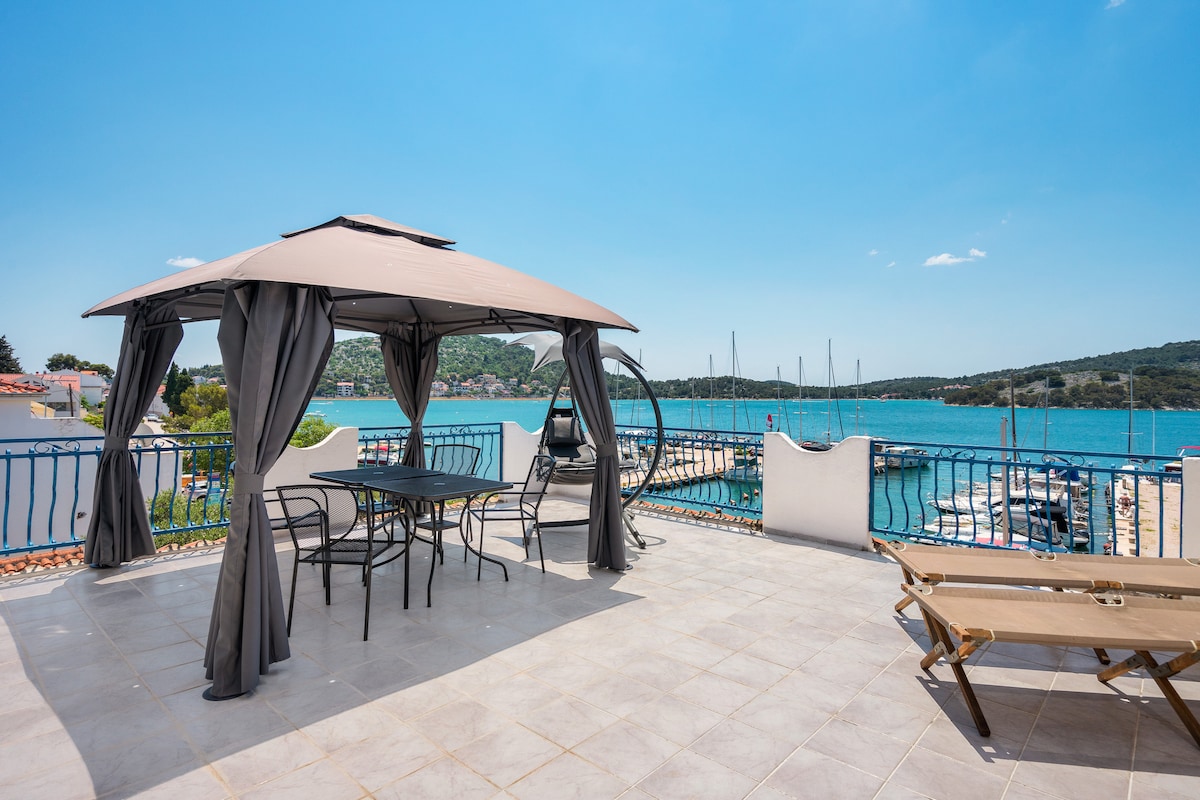 A spacious terrace is enclosed by light grey railings, featuring a gazebo covering a dining table with six chairs. Sun loungers are visible beside the table, offering a view of the nearby marina and bright blue water under a clear sky.