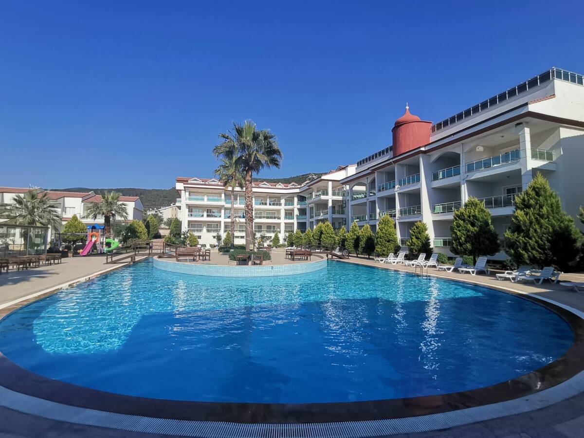 A large outdoor pool is featured in the center of the resort, surrounded by well-landscaped greenery and lounge chairs. Several palm trees enhance the tropical feel, while the Mediterranean-style buildings provide a backdrop against a clear blue sky.
