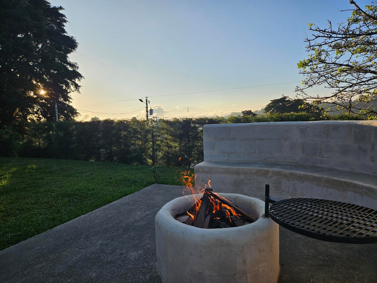 A firewood ring is positioned on a concrete patio, surrounded by a grassy area. A grill grate is visible resting over the circular stone structure. The setting sun casts a warm glow, highlighting trees in the background and a serene landscape beyond.