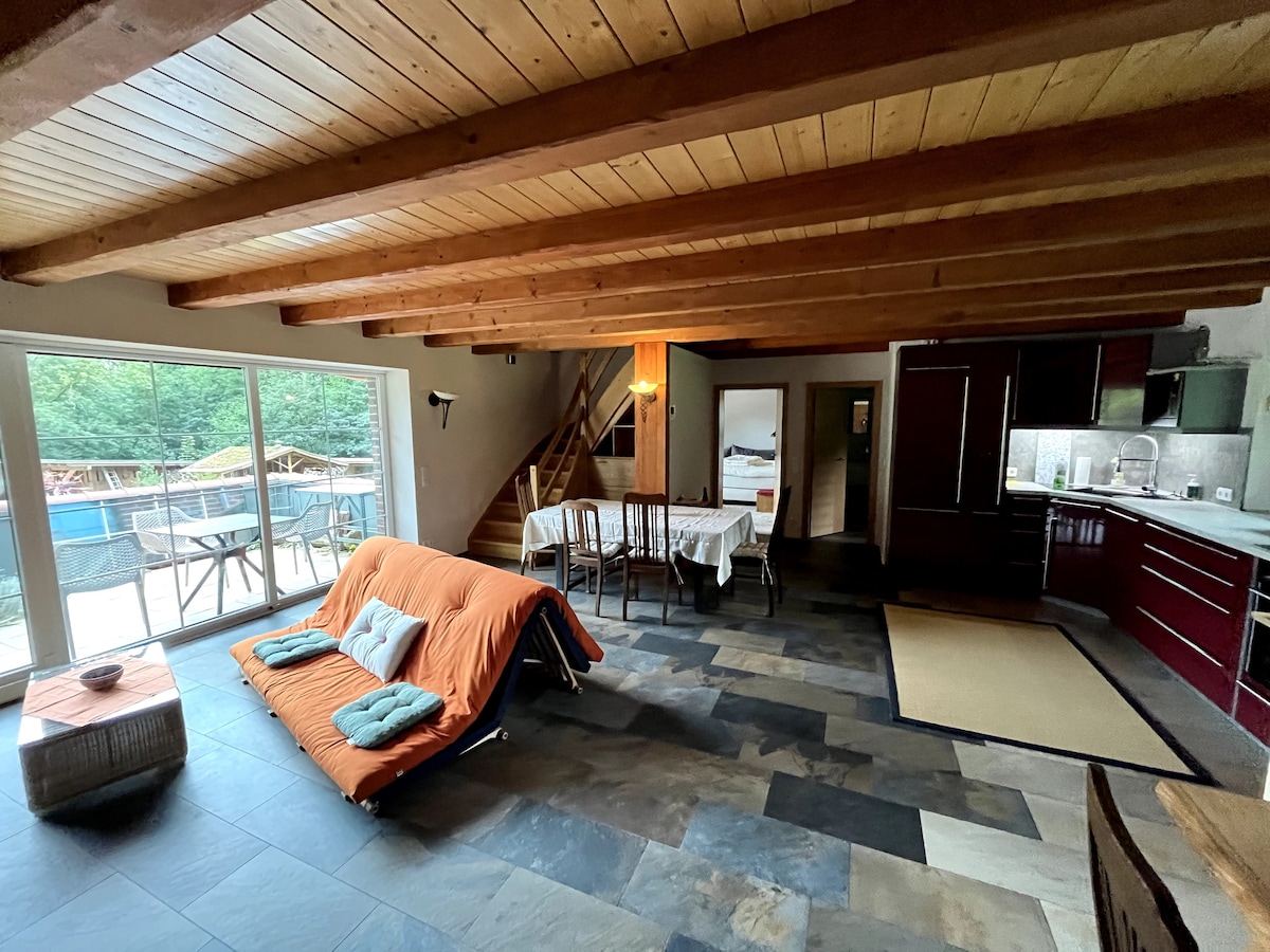 A spacious living area features an orange fold-out sofa and a dining table accommodating four. Large glass doors open to a terrace, while warm wooden beams adorn the ceiling. The kitchen space is equipped with dark cabinetry and modern appliances, enhancing functionality.