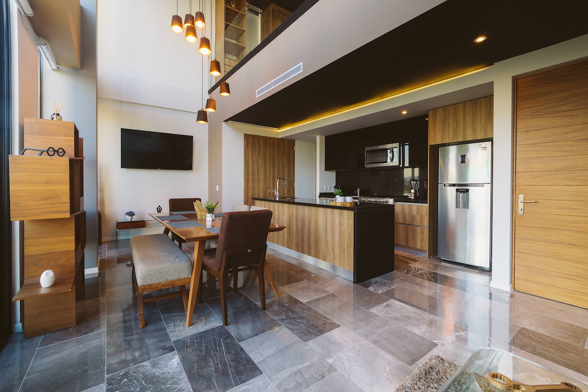 An open-plan living area features a modern kitchen with sleek cabinetry and stainless steel appliances. A wooden dining table is paired with comfortable chairs, while a spacious sofa and wall-mounted television complete the space. Large windows invite natural light, enhancing the minimalistic decor.