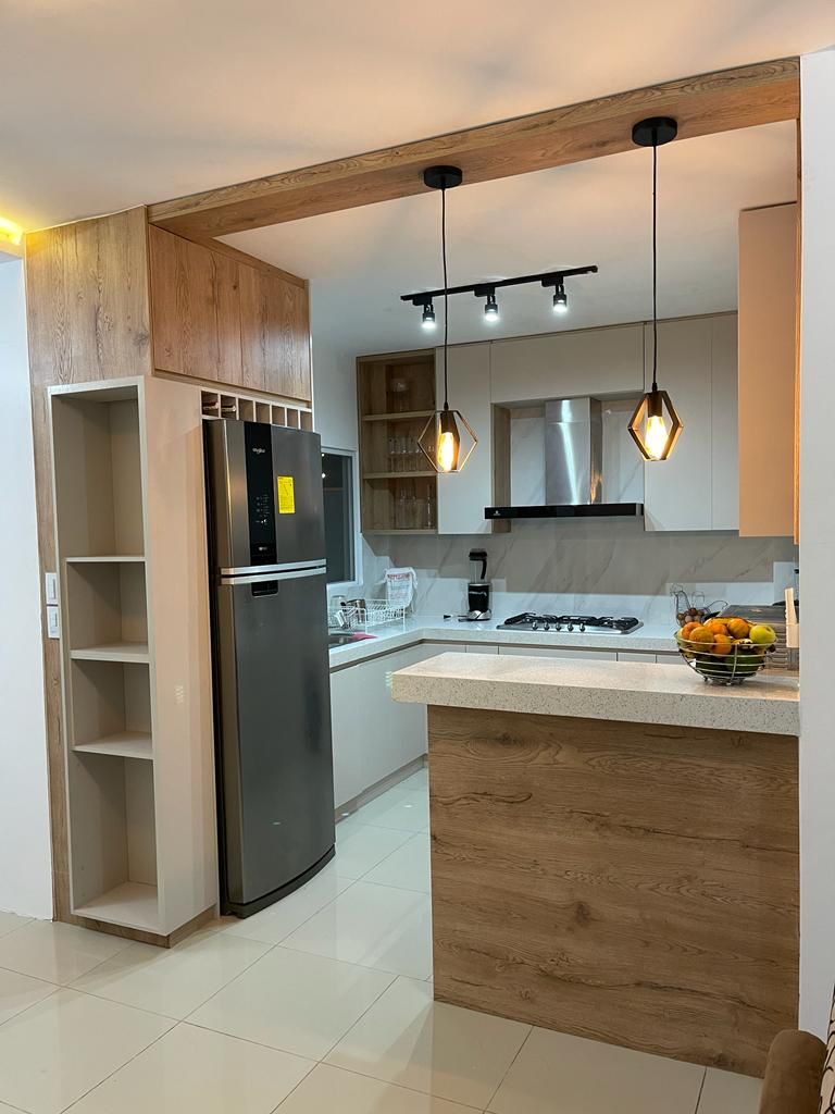The kitchen features modern cabinetry and a functional layout. A stainless steel refrigerator and a built-in oven are visible. Pendant lighting hangs above the counter, which is partially made of light-colored stone. An open shelf displays various kitchen essentials.
