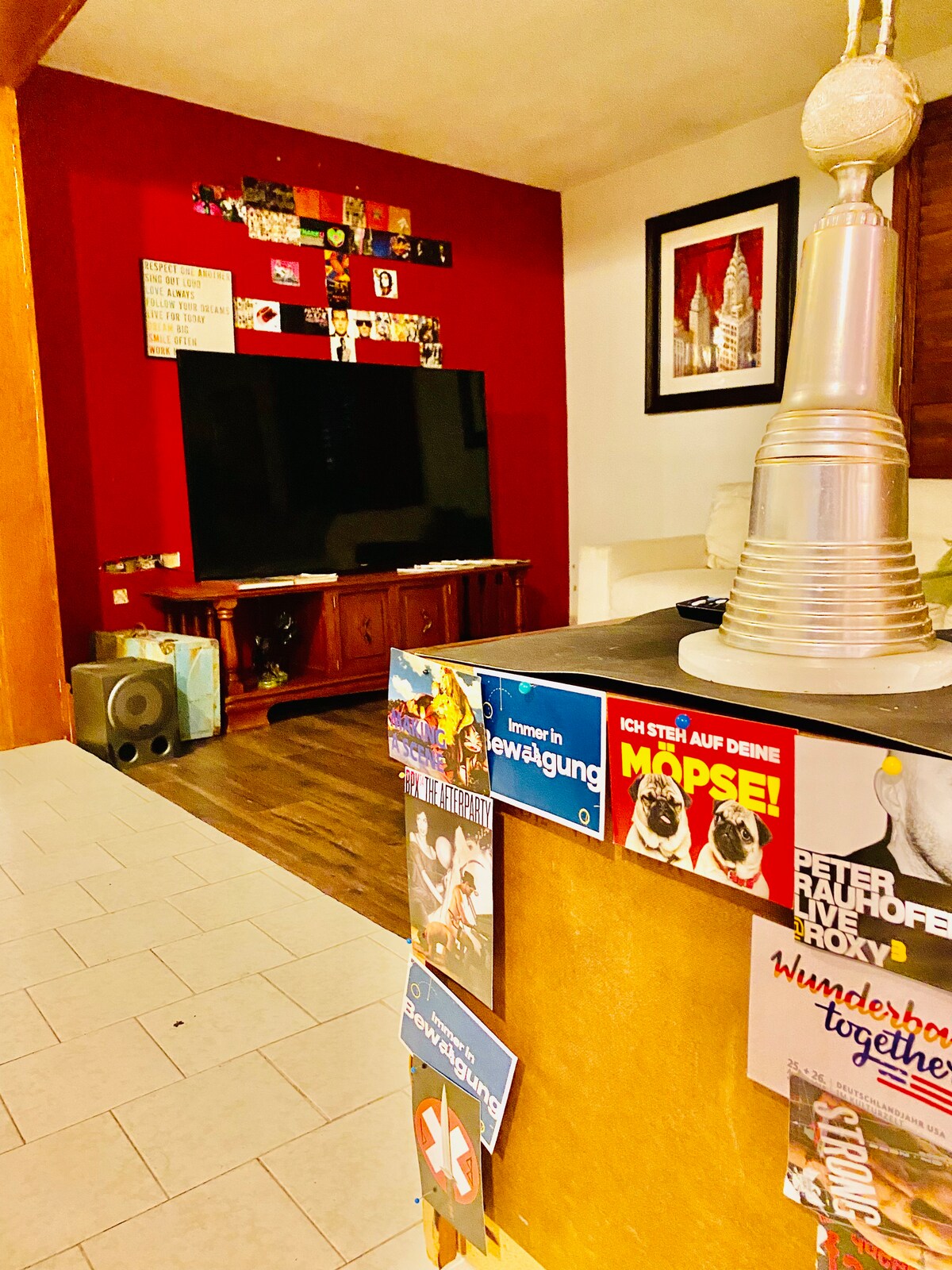 A living area features a large flat-screen TV mounted on a bold red wall. A wooden entertainment unit holds the TV, while colorful magnets decorate a nearby kitchen appliance. Artwork of cityscape enhances the decor, and a light-colored sofa is visible in the background.