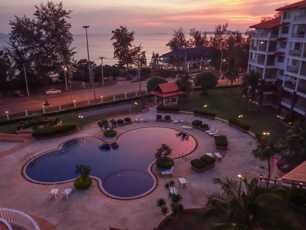 A spacious swimming pool is featured, reflecting the sunset with hues of orange and purple. Surrounding greenery includes well-manicured gardens and decorative shrubs. Several lounge chairs are arranged nearby, contributing to a serene outdoor environment, with views of the ocean in the distance.