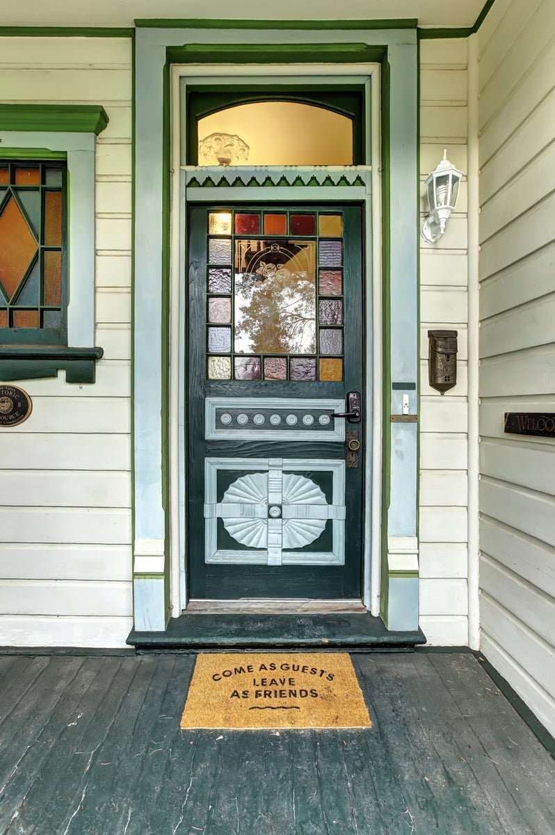 A vintage front door is highlighted, featuring intricate stained glass accents in various colors. A warm welcome mat sits at the threshold, displaying the message 'COME AS GUESTS, LEAVE AS FRIENDS.' The door is framed by a well-maintained exterior and decorative wall features.