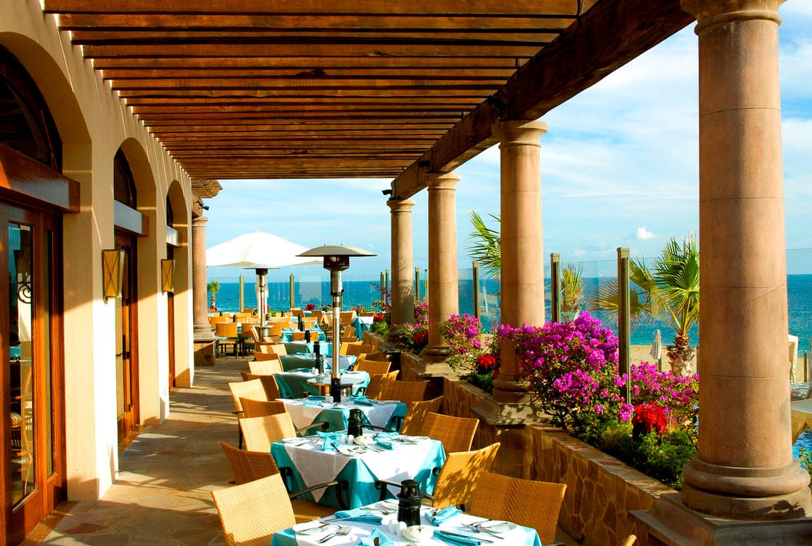 An alfresco dining area is presented with multiple tables set for guests, featuring blue and white tablecloths. The space is framed by sturdy columns and a wooden pergola, while vibrant flowering plants add color to the setting. The ocean can be seen in the background.