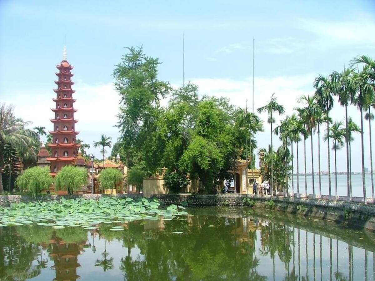 A serene view of a traditional pagoda framed by lush greenery and palm trees. A tranquil pond reflects the vibrant scene, dotted with lotus leaves. The peaceful atmosphere is enhanced by the nearby West Lake, creating a harmonious blend of nature and culture.
