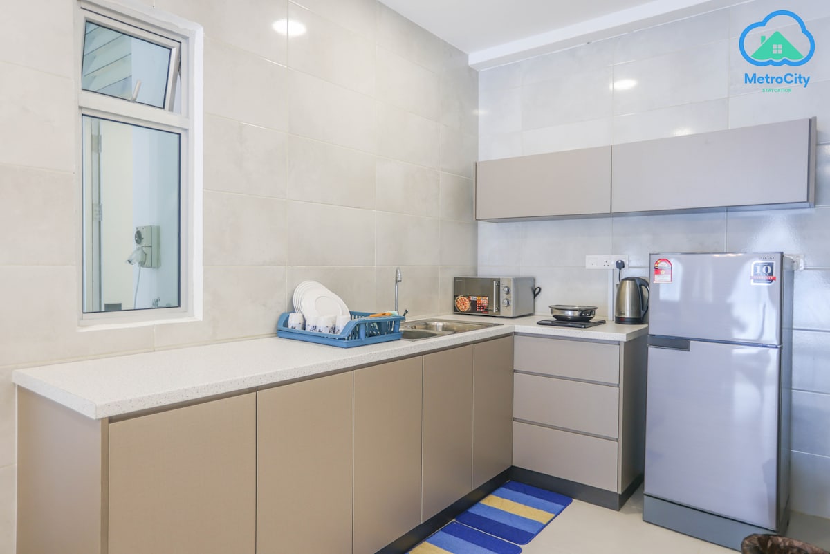 A modern kitchen features light grey cabinetry and a sleek countertop. A window allows natural light to brighten the space. Kitchen appliances include a refrigerator, microwave, and kettle. A dish rack holds plates, and a striped rug adds a pop of color to the floor.