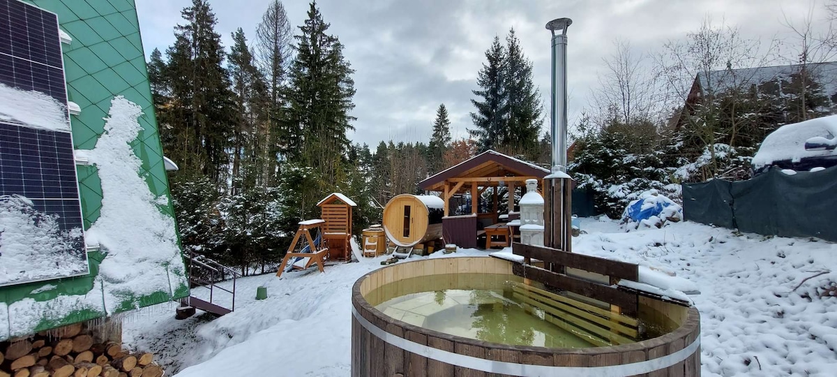 An outdoor area features a wooden hot tub surrounded by a snowy landscape. Nearby, a wooden gazebo and play structure are visible, along with stacks of firewood. Various trees create a natural backdrop, adding to the peaceful ambiance of the setting.