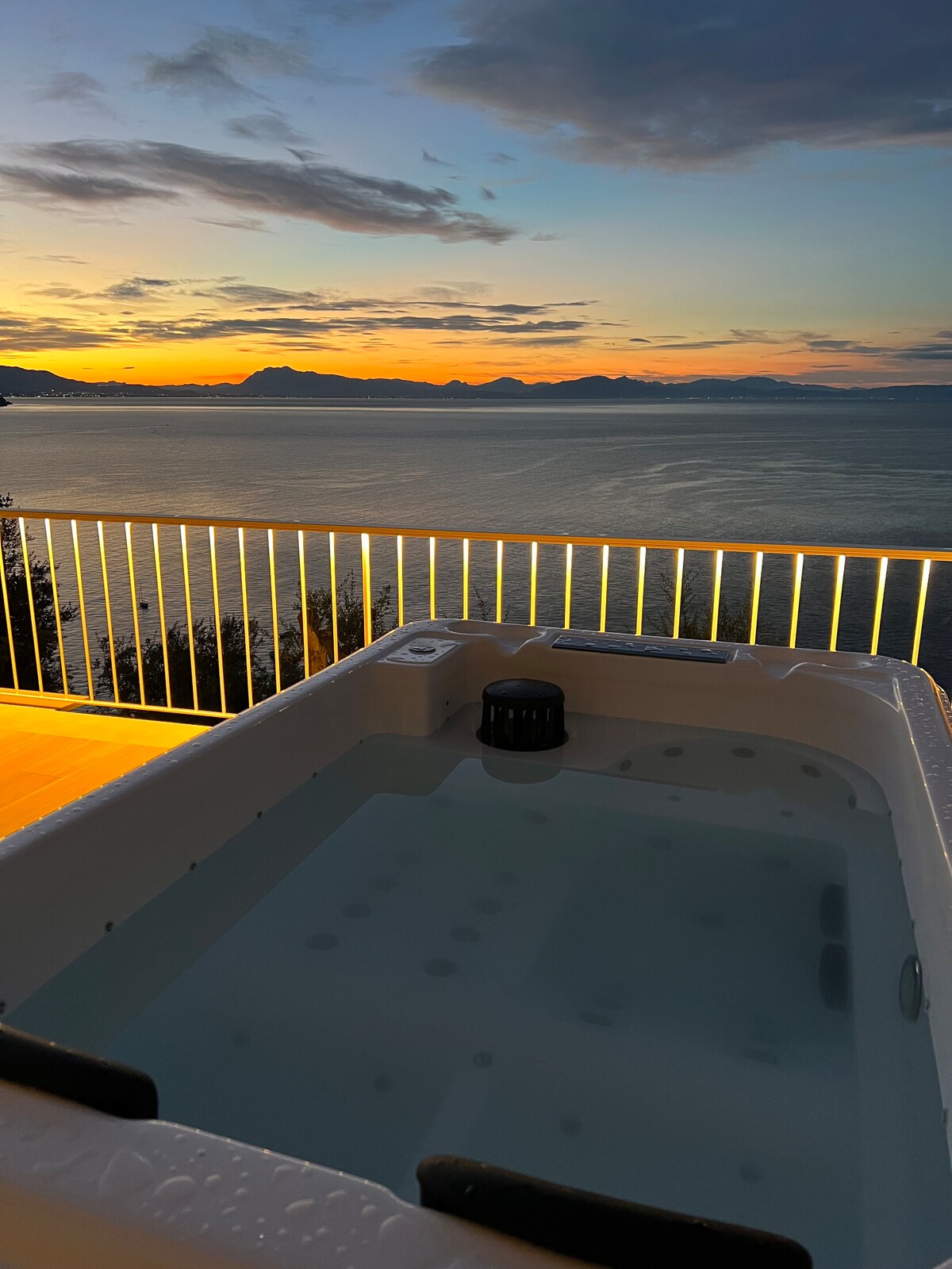 A hot tub is positioned on a terrace overlooking the sea, with a panoramic view of distant mountains at sunset. The sky is filled with warm colors, and the water in the tub is calm, reflecting the tranquil atmosphere.