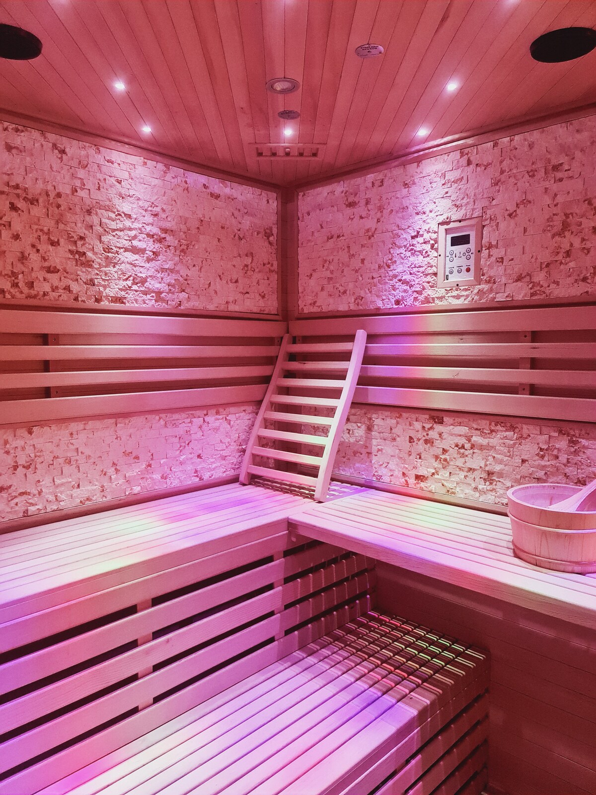 A private sauna is presented with warm wooden benches and an elegant design. Soft lighting illuminates the space, creating a relaxing atmosphere. A wooden bucket rests on the floor, and a control panel is visible on the wall.