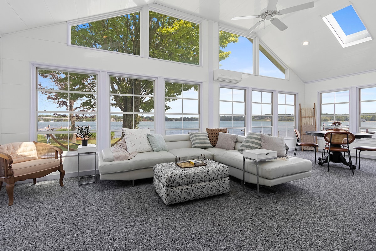 A three-season porch features large windows that allow for ample natural light and provide views of the water. Comfortable sectional seating is positioned around a low ottoman, while a dining table and chairs are visible in the background. A ceiling fan enhances airflow.
