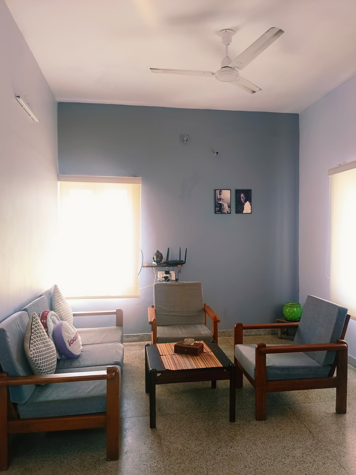 The living room offers a cozy seating arrangement featuring two armchairs and a two-seater sofa. A wooden coffee table is positioned centrally. Natural light enters through two windows, complemented by light blue walls. A ceiling fan is visible overhead.