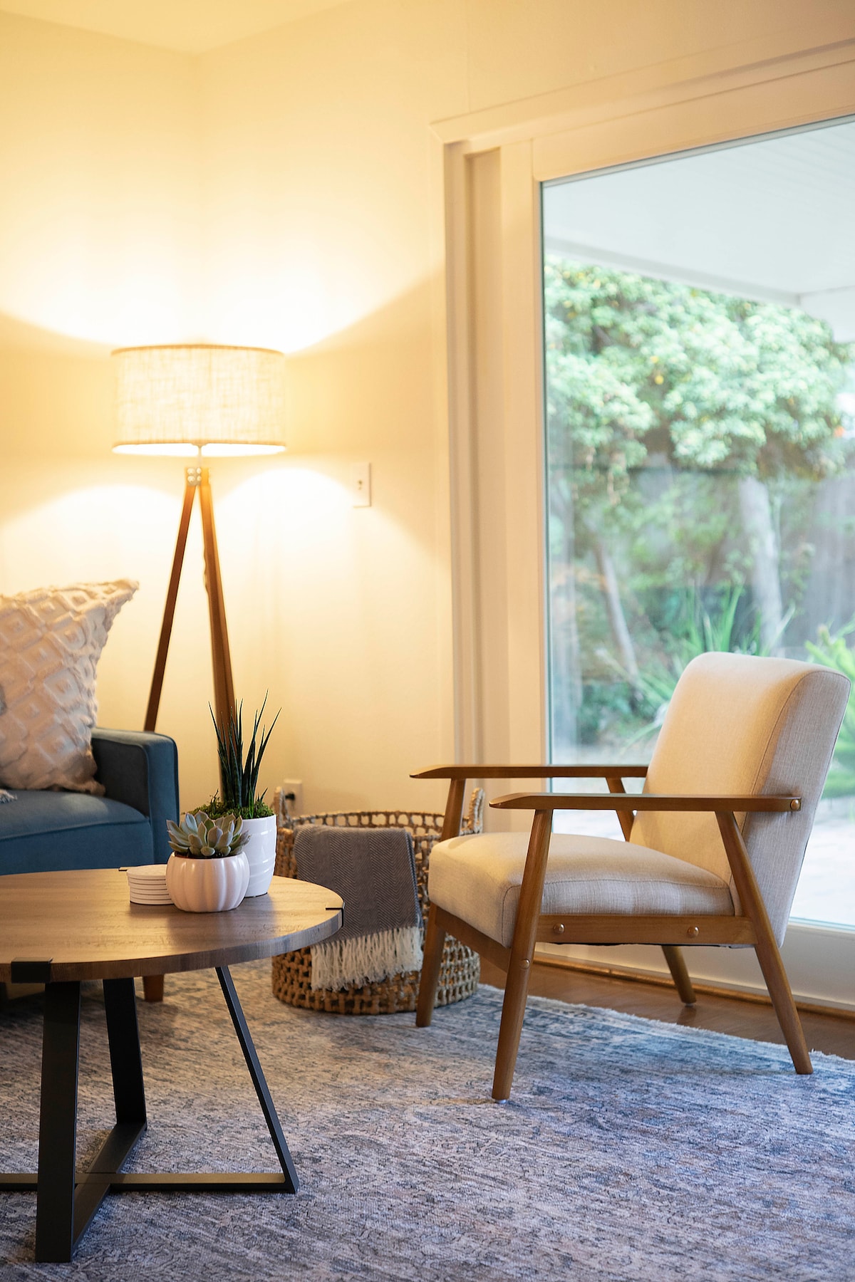 A cozy corner features a light wood chair beside a round coffee table. A soft light illuminates the space, revealing a small potted plant on the table. Large glass doors offer a view of a lush, green outdoor area, enhancing the welcoming atmosphere.