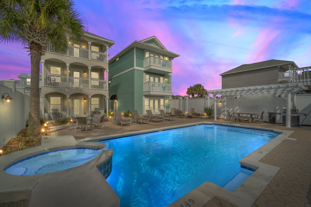 A serene outdoor space features a large pool surrounded by lounge chairs, illuminated by soft lighting at dusk. The multi-story beach house stands nearby, showcasing balconies that overlook the pool, with palm trees adding a touch of greenery to the setting.