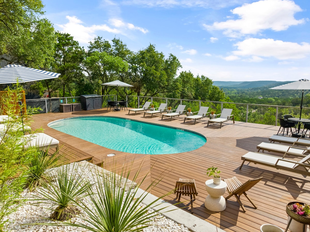 A serene outdoor area features a smooth pool surrounded by sun loungers on a wooden deck. A dining area is visible nearby, shaded by large umbrellas, while lush trees frame a distant view of rolling hills under a clear blue sky.