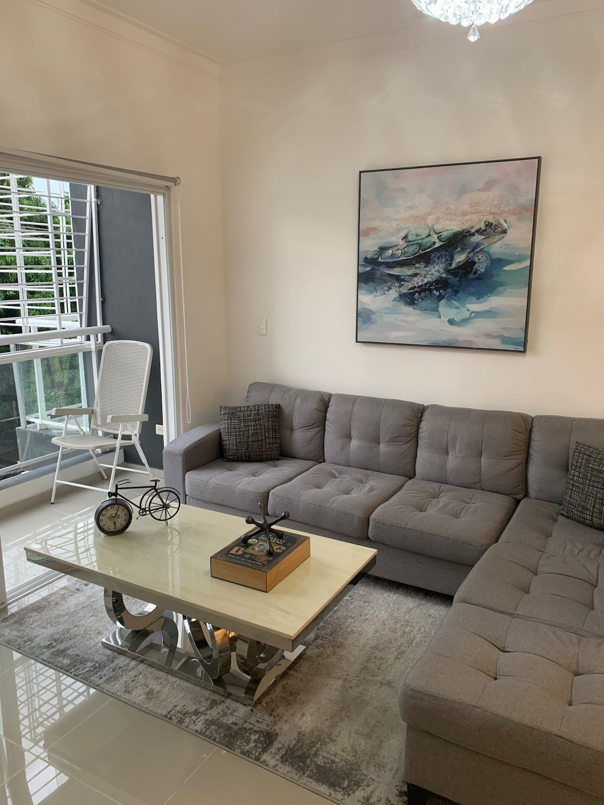 A modern seating area is presented with a gray sectional sofa, featuring decorative pillows. A glass coffee table occupies the center, accompanied by a decorative clock and a small bicycle model. A large colorful painting adorns the wall, while natural light is filtered through the nearby window.