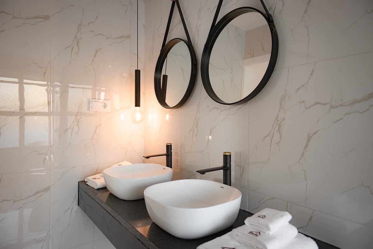 A modern bathroom features two round sinks positioned on a dark countertop. Each sink is accompanied by a stylish black faucet. Above them, two circular mirrors are suspended by black geometric frames. Warm lighting is provided by pendant lights, and neatly arranged towels are visible nearby.