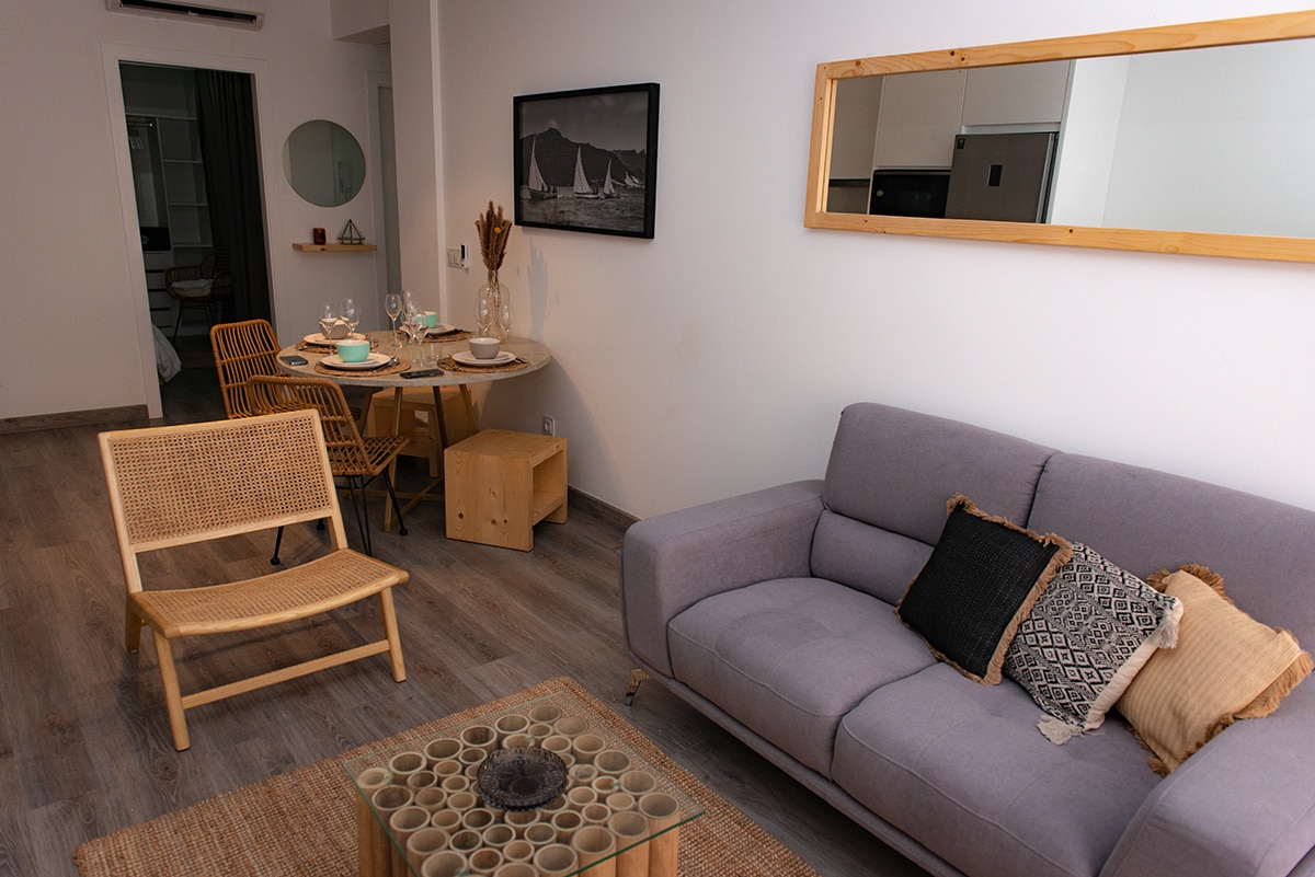 A modern living area is showcased with a light gray sofa, accompanied by a natural wood chair. A round dining table is set for four, with decorative items and tableware. A large mirror enhances the space, and light wood flooring complements the neutral colors.