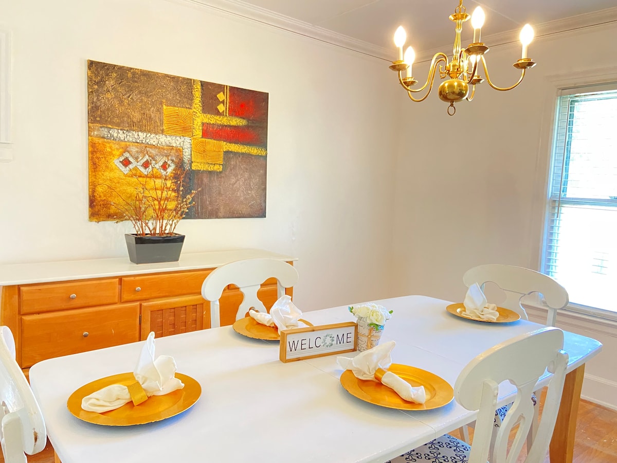A bright dining area features a white table set for four, with gold plates and neatly folded napkins. A decorative welcome sign sits at the center. A vibrant abstract artwork adorns the wall behind, complemented by a chandelier that provides gentle lighting.
