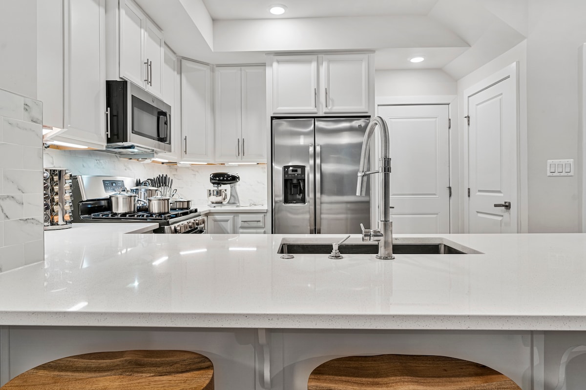 A modern kitchen features sleek white cabinetry and a large island with a quartz countertop. Stainless steel appliances, including a microwave and refrigerator, are visible. Two wooden bar stools are positioned at the island, providing seating at the breakfast area.