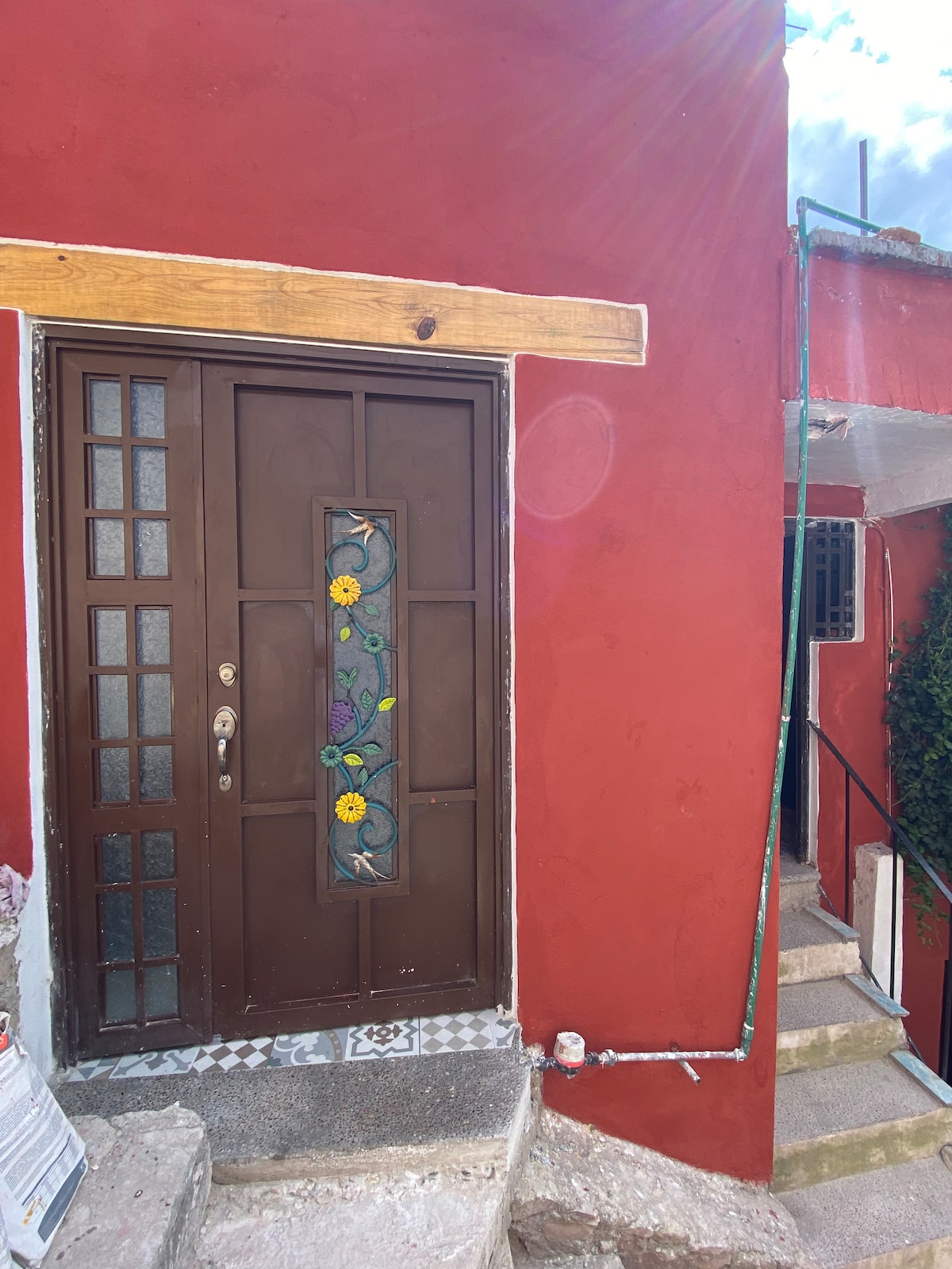 The entrance displays a brown door featuring a decorative glass panel with floral designs. The wall is painted in a vibrant red, and a set of stone steps leads up beside the entrance, flanked by greenery.