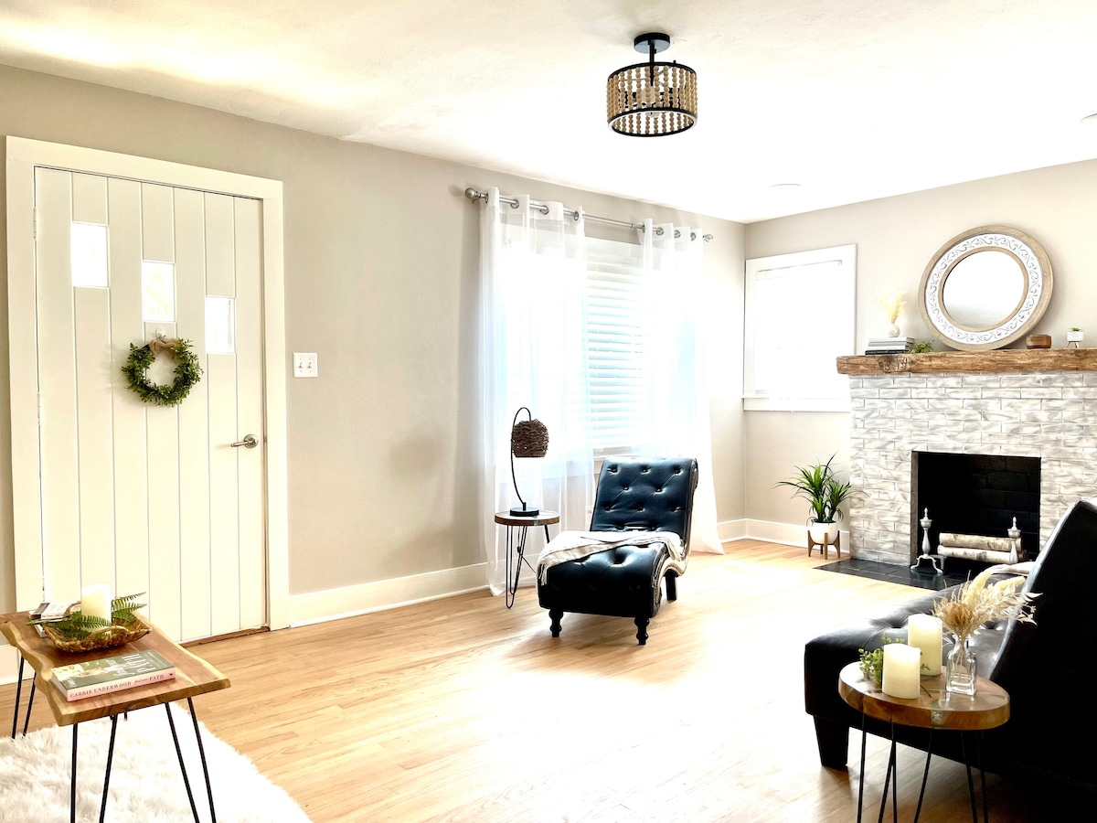 A light-filled living room features a stylish fireplace with a stone surround and a large mirror above it. An upholstered chair and a small side table are positioned near a doorway, while sheer curtains allow natural light to filter in. Indoor plants add a touch of greenery.