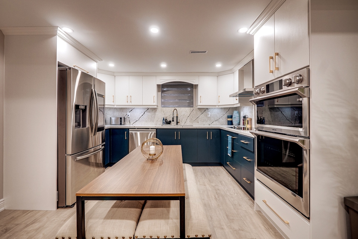 The kitchen features modern cabinetry in blue and white, with stainless steel appliances including a refrigerator and ovens. A spacious island is set with a wooden dining table and decorative centerpiece, complementing the sleek countertop and tile backsplash.
