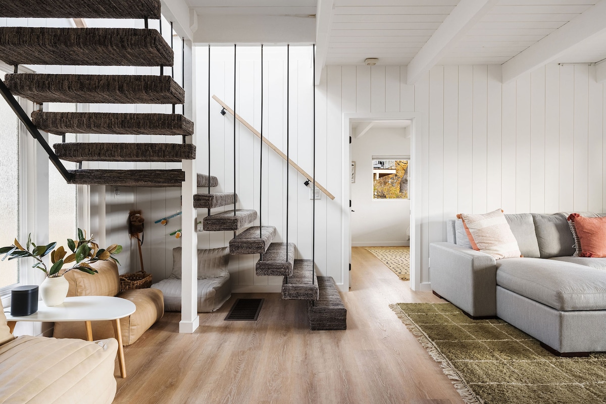 The modern interior features an open staircase with textured steps leading to an upper level. A light-colored sectional sofa is seen in the living area, complemented by a decorative rug. Potted plants add a touch of greenery to the inviting space.