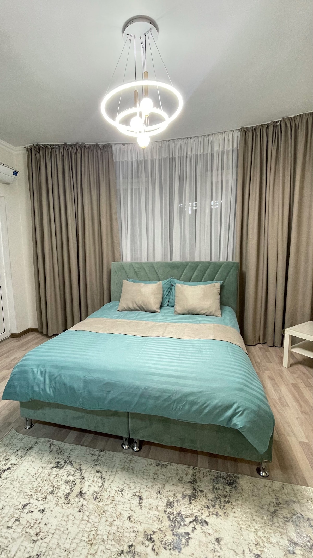 A sleeping area is presented with a green bed featuring a tufted headboard and light textiles. Two decorative pillows are centered on the bed. Sheer curtains frame a window, and a simple light fixture hangs above, enhancing the calm ambiance.
