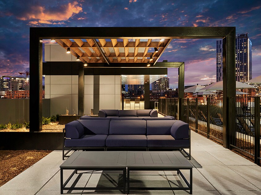 An outdoor rooftop area features a comfortable lounge setup with a dark blue sectional sofa and a matching coffee table. The wooden pergola provides shade while city skyline views are visible in the background, highlighted by an evening sky.
