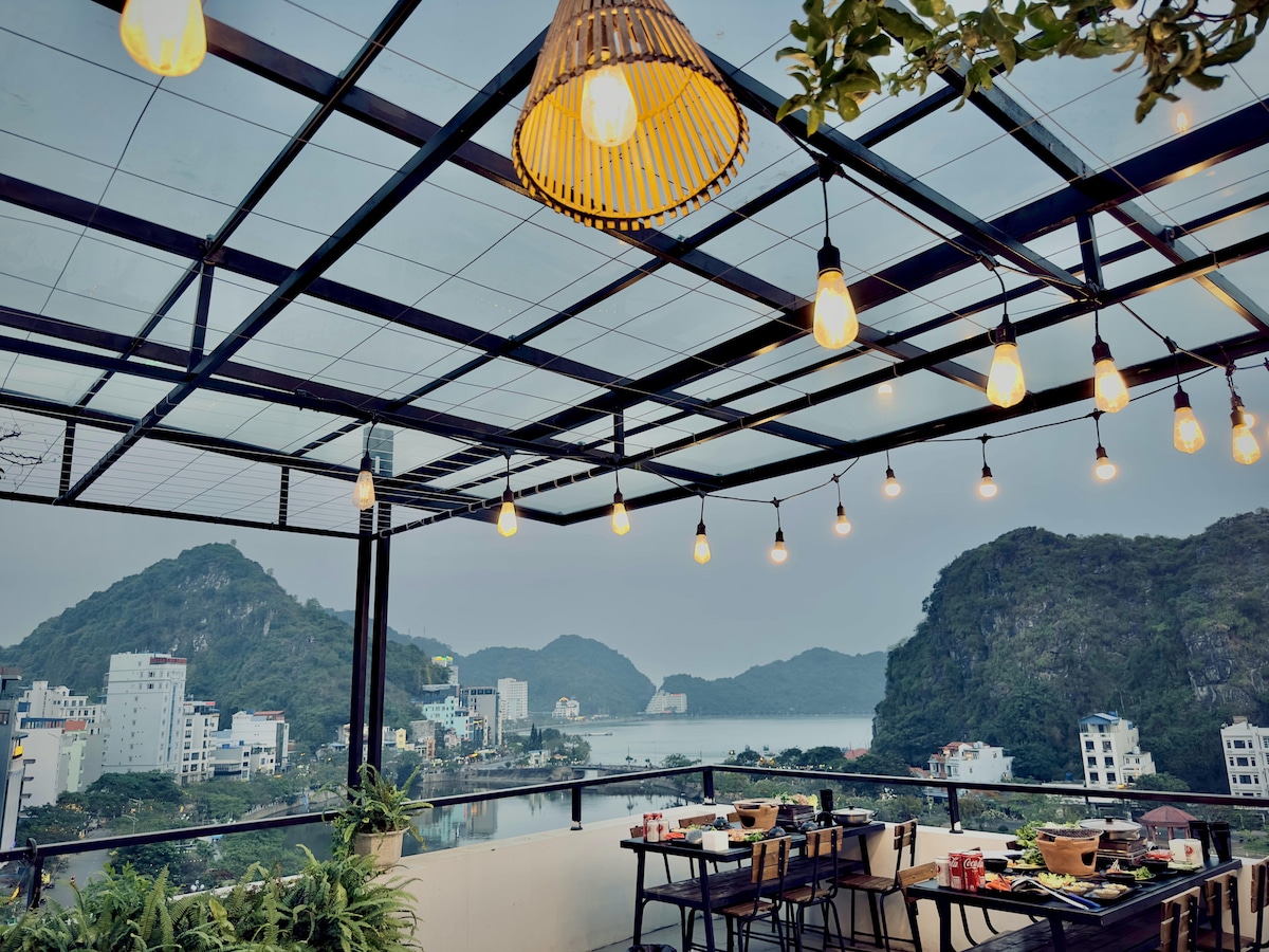 An outdoor terrace features a modern design with a transparent roof showcasing a series of light fixtures hanging above. Tables are arranged with dining items, providing picturesque views of the bay and surrounding hills in the background.