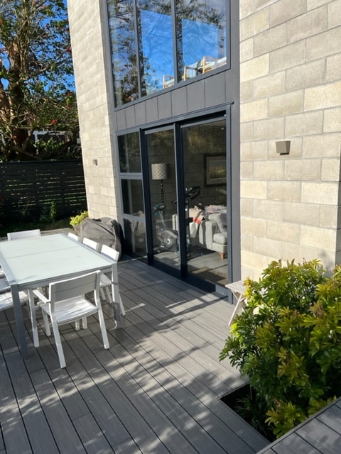A spacious outdoor deck features a white dining table surrounded by matching chairs. Large glass doors provide a view into the interior space, enhancing the connection between indoors and outdoors. Green plants line the area, contributing to a tranquil setting.