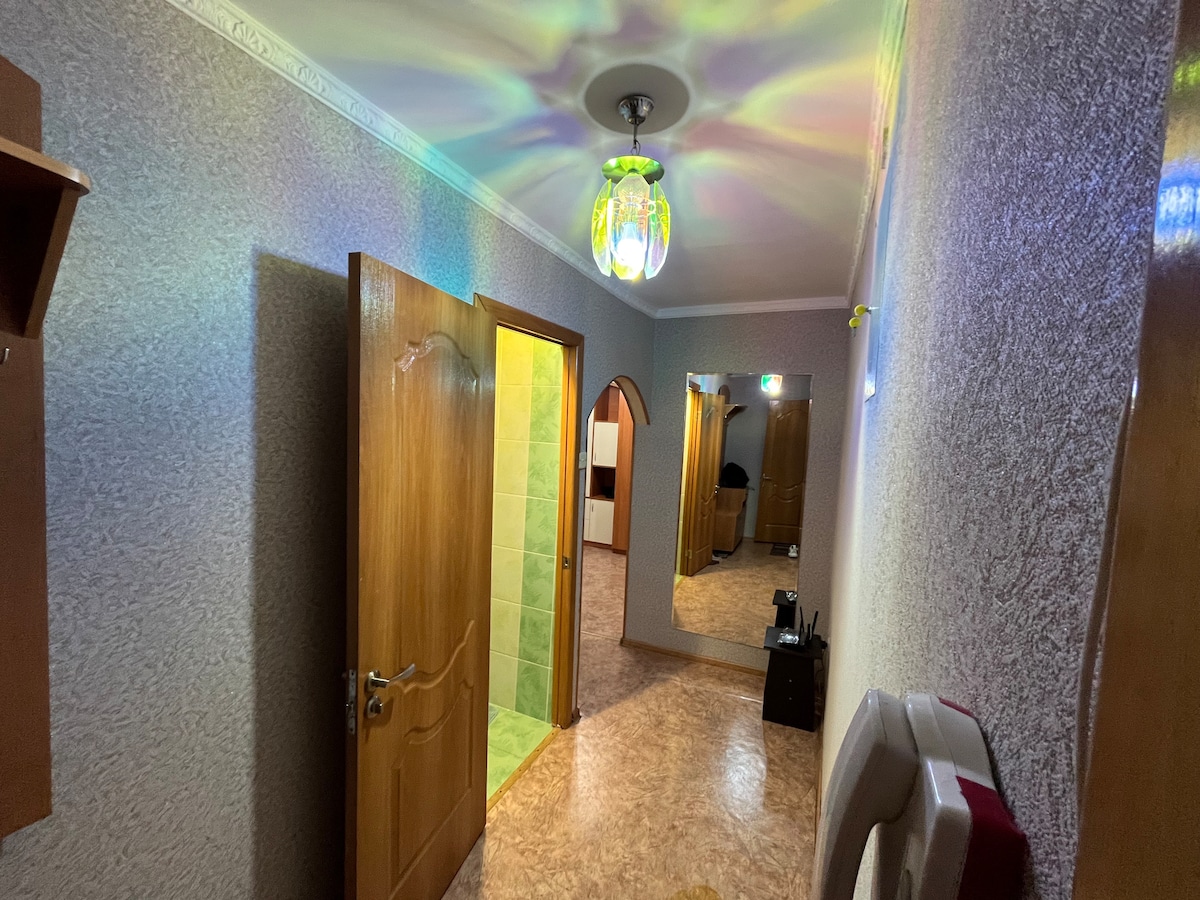 A hallway is shown, featuring wooden doors leading to various rooms. The walls have a textured finish, complemented by a colorful light fixture that casts soft reflections. A mirror stands prominently on one side, and a small table with a lamp is visible.