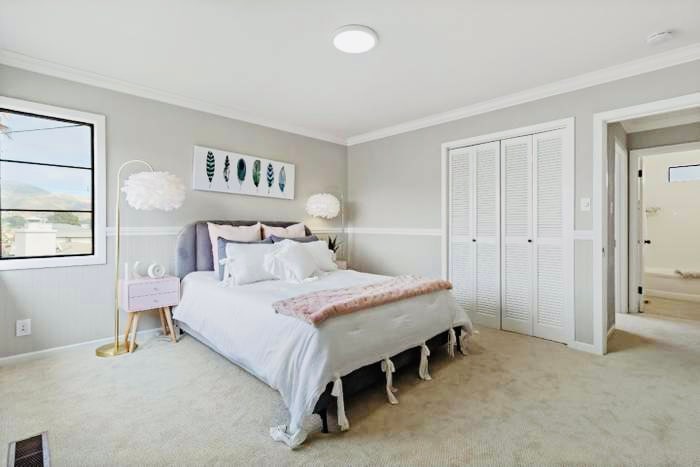 A well-appointed bedroom features a spacious bed with light bedding and decorative pillows. Two hanging light fixtures add visual interest. A bedside table holds a lamp, while a large window offers natural light. Closet doors with a louvered design provide additional storage.