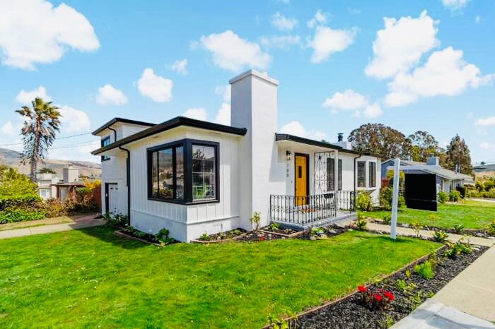 A charming single-story home is framed by lush green grass and colorful landscaping. Large windows allow natural light to fill the space, while a welcoming front door stands out against the white exterior. A decorative chimney adds character to the roofline.